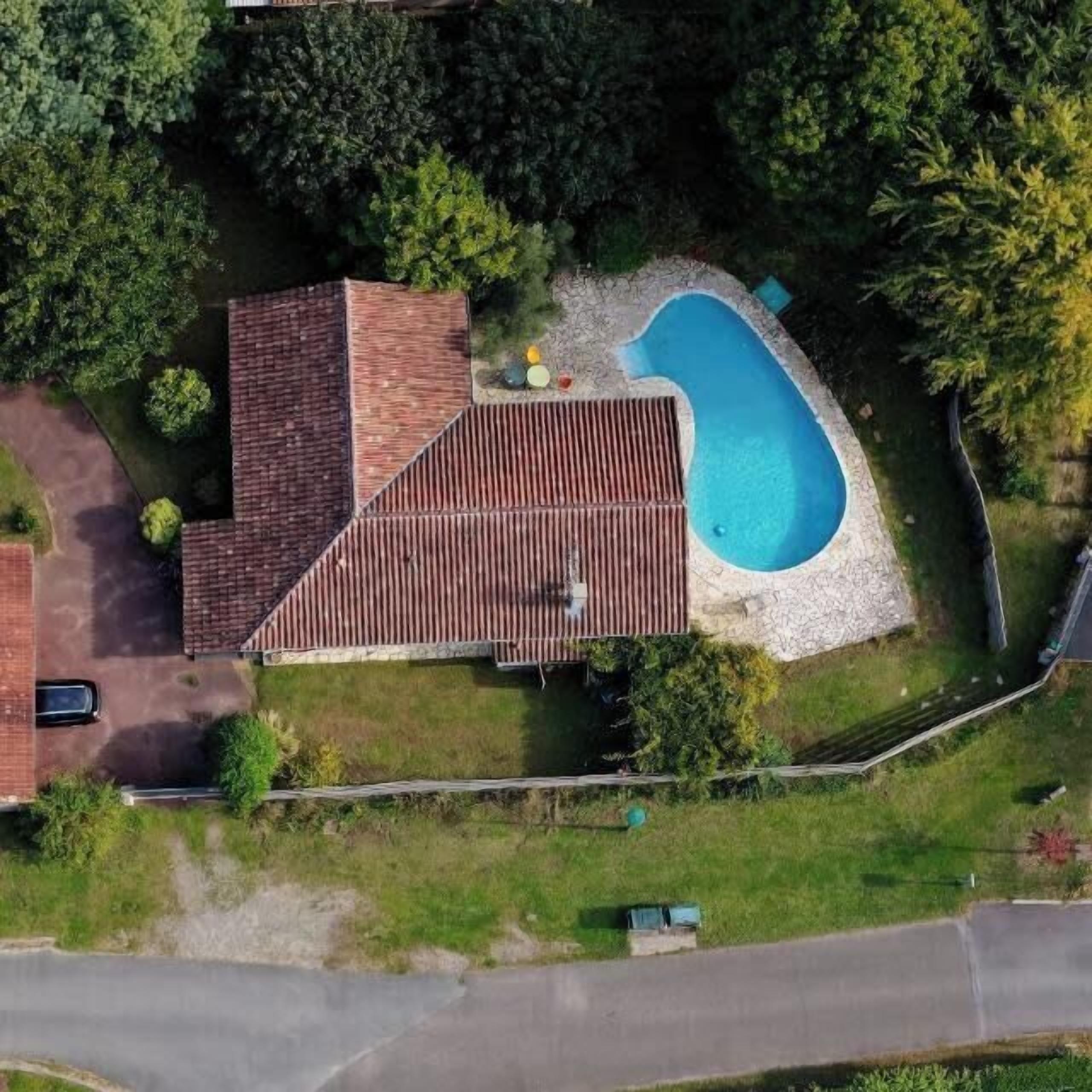 An aerial view of a house with a swimming pool, surrounded by trees and a paved driveway.