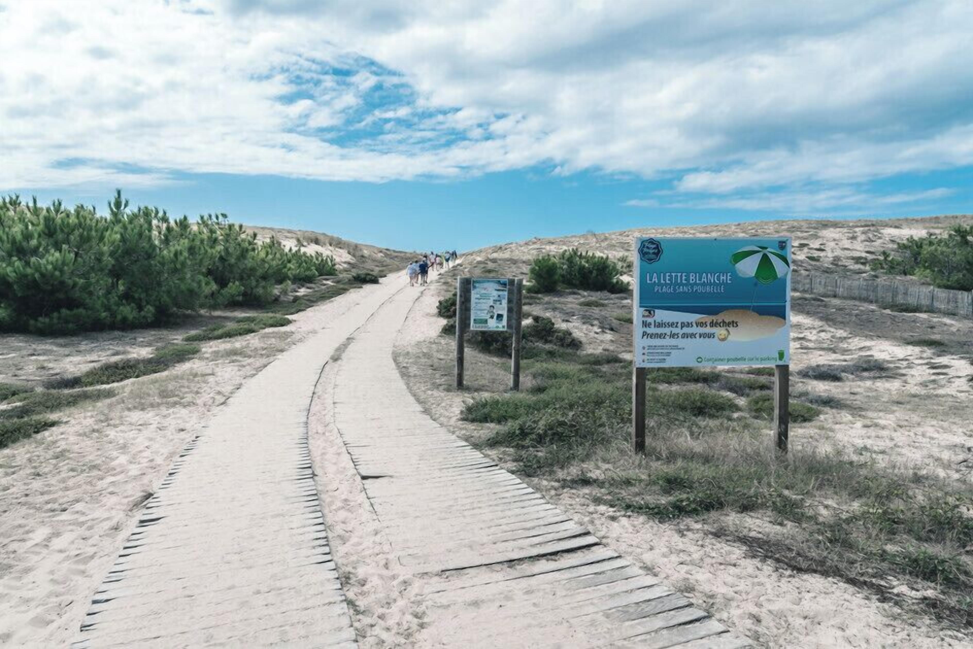 A sandy path leading to a beach area with signs indicating 'La Litte Blanche' and 'Plages Sans Poubelle'.