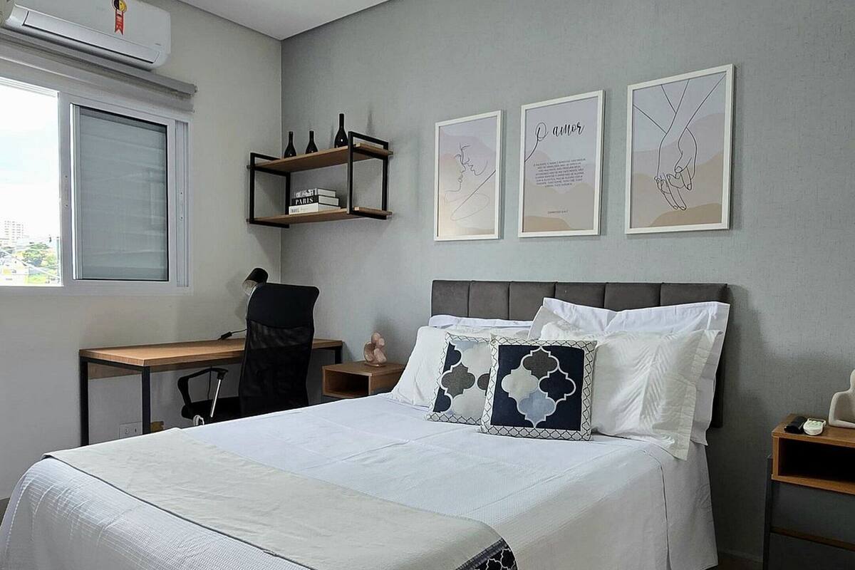 A bedroom with a bed, a desk, a chair, a nightstand, and a wall-mounted shelf.