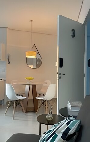 A modern living room with a dining area, a round table, two white chairs, a sofa, a small side table, and a hanging light fixture.