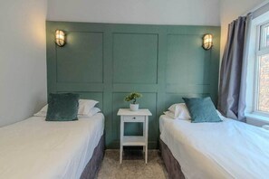 A room with two beds, a small white nightstand with a plant, and wall-mounted lights.