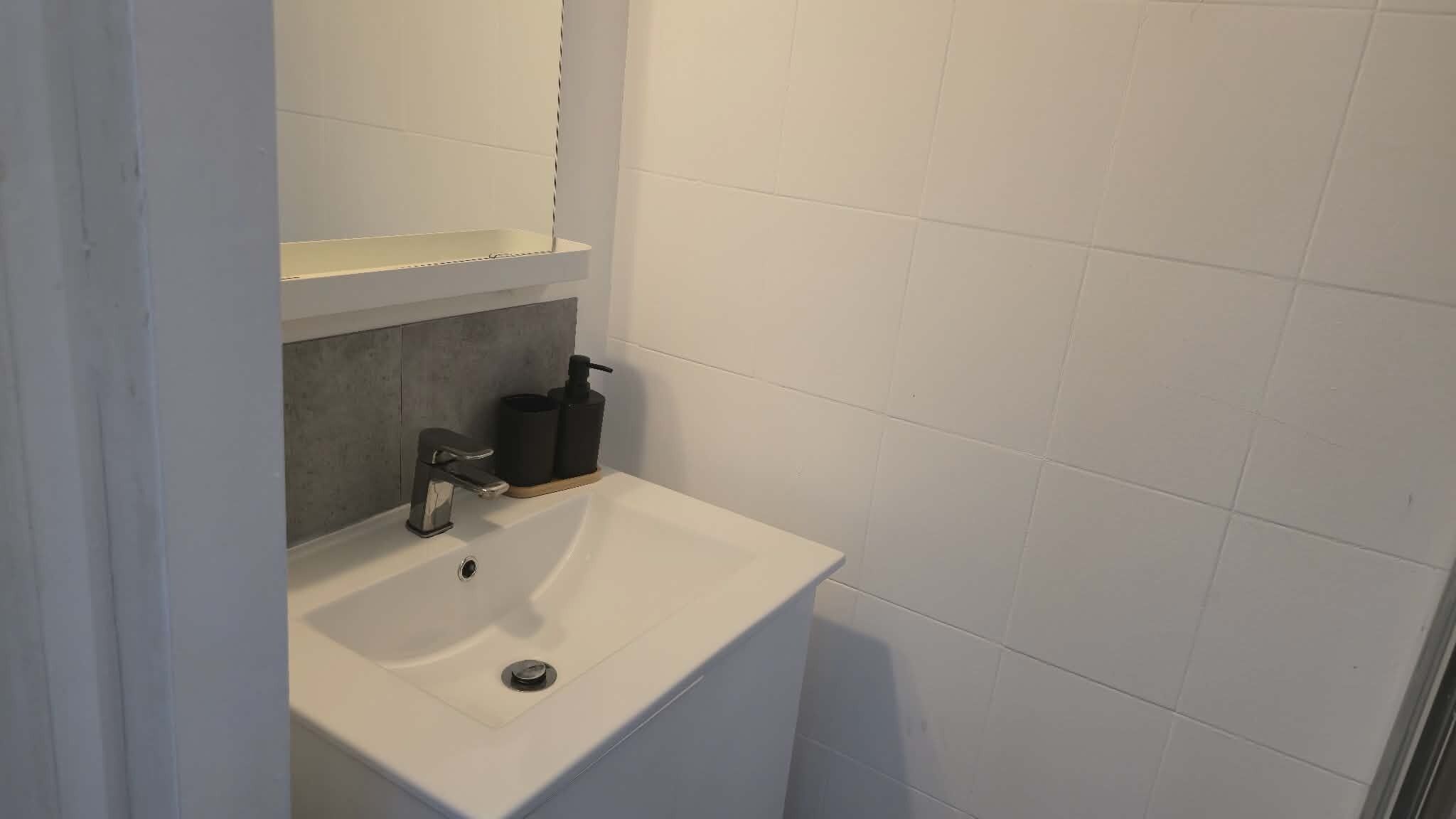 Un baño con un lavamanos blanco, un espejo encima y un dispensador de jabón negro sobre la encimera.