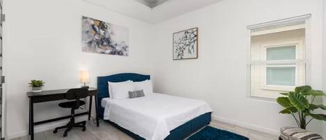 A bedroom with a bed, desk, chair, ceiling fan, artwork, window, and potted plant.