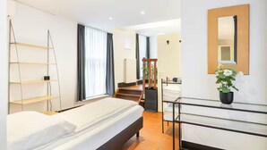 Habitación de hotel con cama, escritorio y silla, espejo, estante y una escalera que lleva a otra habitación.