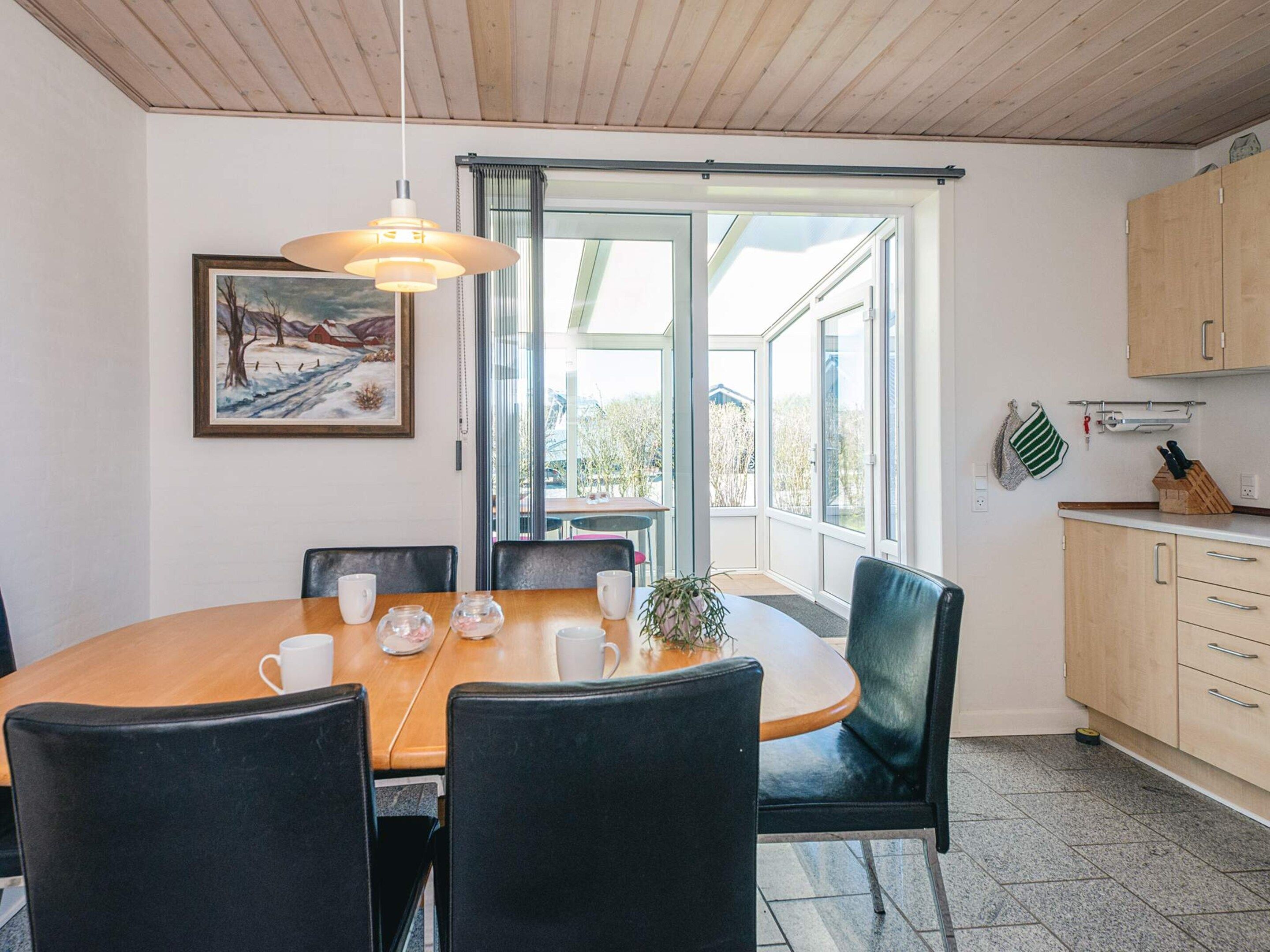 A dining area with a wooden table and chairs, a painting on the wall, and a view through large windows.