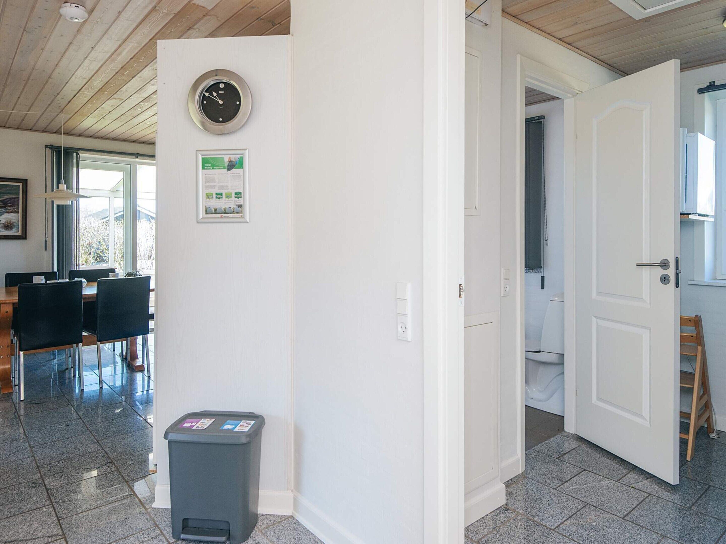 A room with a wooden ceiling, a clock on the wall, a trash bin, and a door leading to another room.