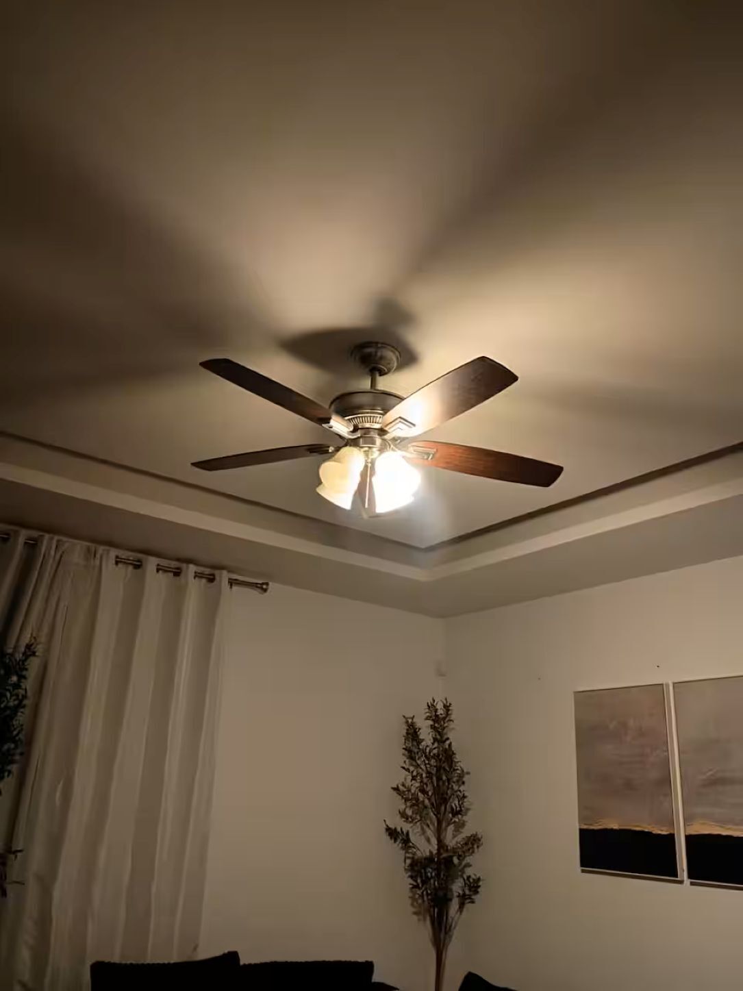 Ein Deckenventilator mit vier Flügeln und einer Leuchte in einem Zimmer mit weißer Wand, einem Fenster mit Vorhängen und einer Pflanze.
