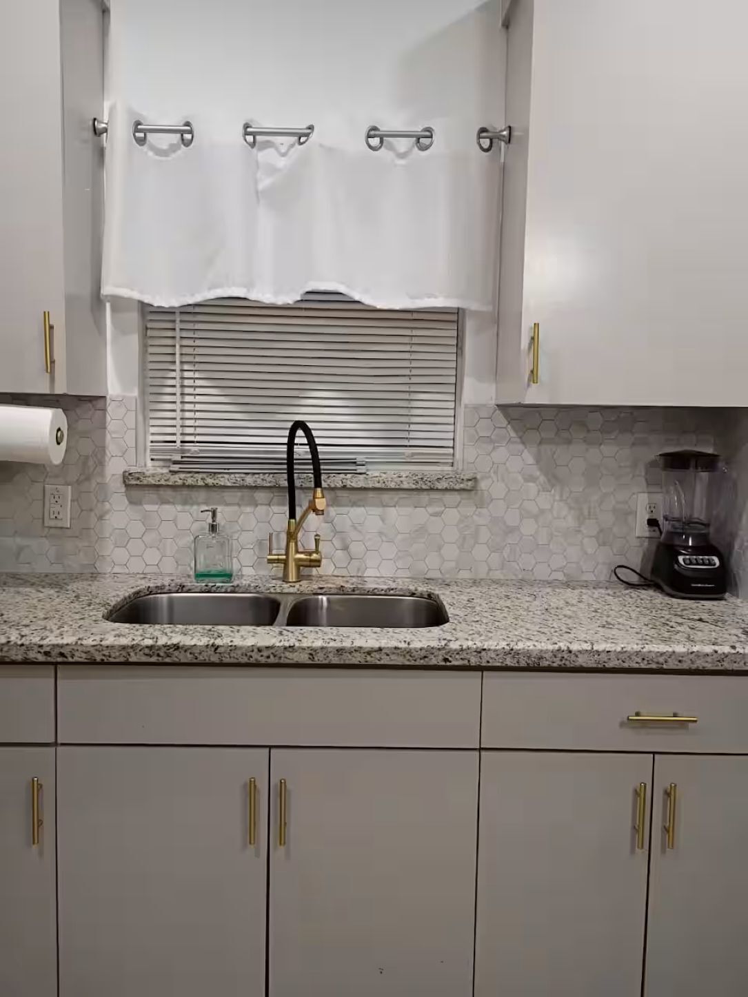 A kitchen with a stainless steel sink, a faucet, a window with blinds, and a blender on the counter.