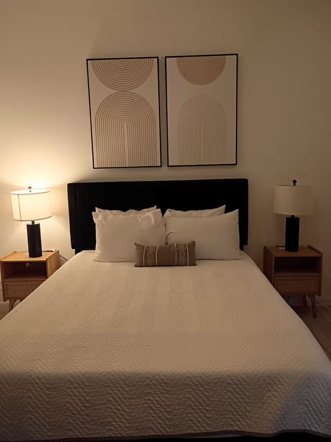 A neatly made bed with a white duvet, two pillows, and a decorative pillow, flanked by two bedside tables with lamps, in a room with a dark headboard and two wall-mounted artworks.