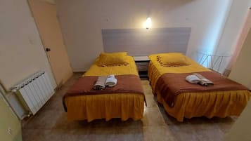 A room with two beds, each with a brown bedspread and a rolled towel. There is a radiator on the left wall and a light fixture above the beds.
