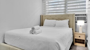 A neatly made bed with rolled towels, a bedside lamp, and a window with blinds. - Causeway House | Coastal 2BR Stay (Bay Harbor Islands)