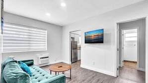 A modern living room with a sofa, a round table, a flat-screen TV, and a view of a sunset through a window. - Causeway House | Coastal 2BR Stay (Bay Harbor Islands)
