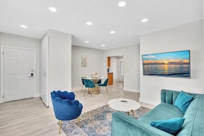 A modern living room with a teal sofa, a blue armchair, a dining area with a round table, and a large painting of a sunset. - Airy 2BR in Bay Harbor Islands close to beach, shopping, and dining (Bay Harbor Islands)