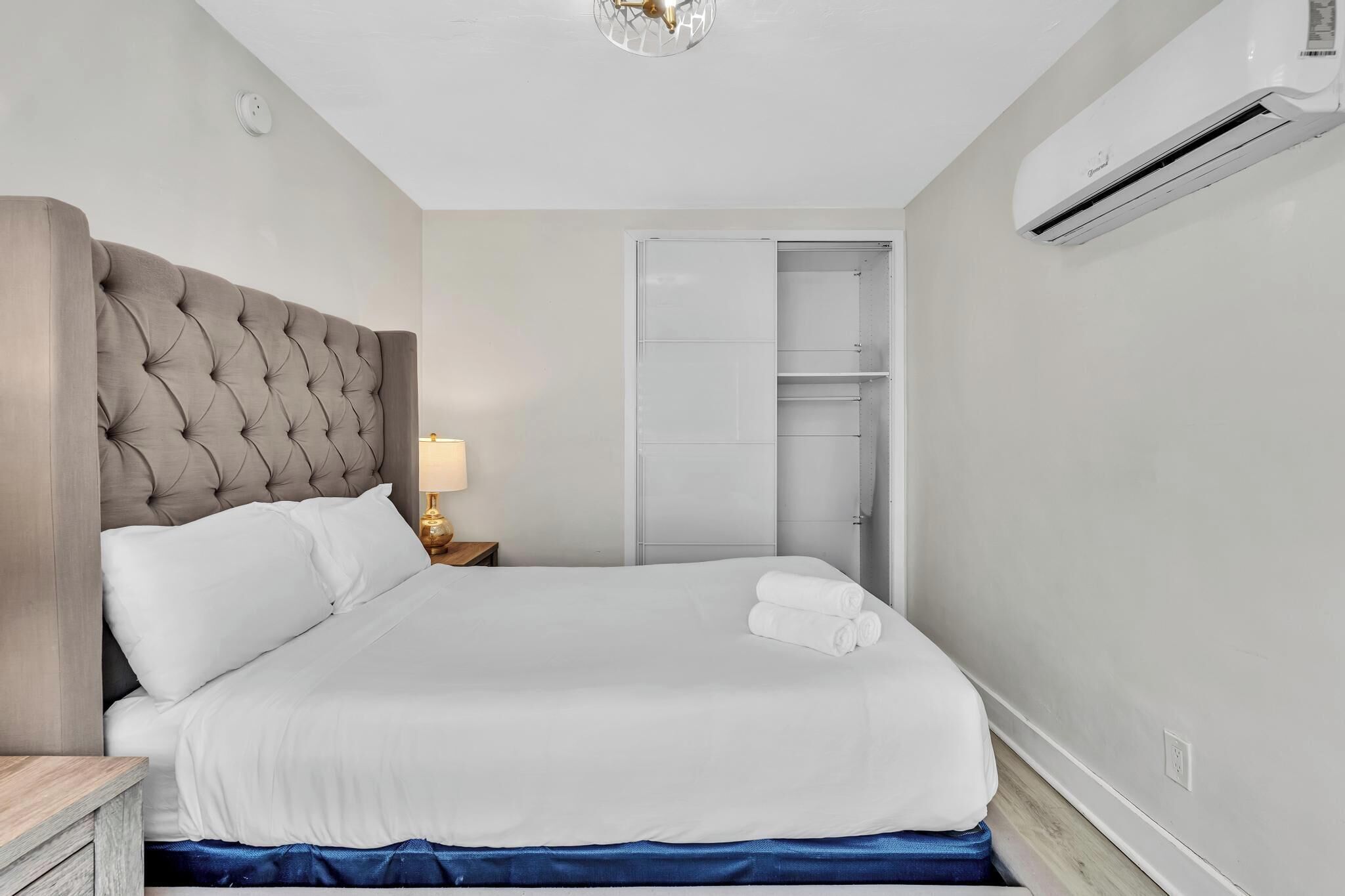 A neatly made bed with a tufted headboard, a bedside lamp, and an air conditioning unit mounted on the wall.