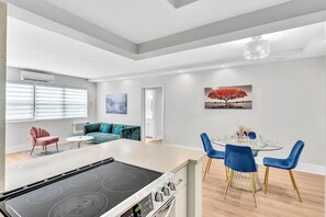 A modern kitchen with a central island, a dining area with blue chairs, and a living space with a sofa and a painting. - Old Bay Vista | Stylish 1BR Getaway (Bay Harbor Islands)