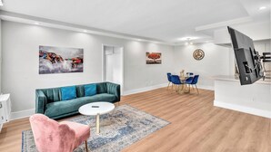 A modern living room with a green sofa, a pink chair, a round coffee table, and a blue rug. There is a dining area with blue chairs and a table, and a TV mounted on the wall. - Old Bay Vista | Stylish 1BR Getaway (Bay Harbor Islands)