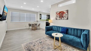 Un salon moderne avec un canapé bleu, une petite table ronde, un téléviseur et un coin repas avec des chaises.