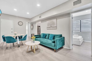 Ein modernes Wohnzimmer mit einem türkisfarbenen Sofa, einem runden Couchtisch und türkisfarbenen Sesseln. Im hinteren Bereich befindet sich ein Essbereich mit einem weißen Tisch und Stühlen. Ein weißes Sofa ist im Hintergrund zu sehen.
