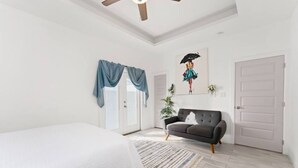 Une chambre à coucher avec un lit, un canapé sombre, un ventilateur de plafond, une fenêtre avec des rideaux bleus et un tableau au mur.