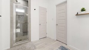 Un couloir avec une porte de douche en verre, deux portes blanches et une petite étagère sur laquelle est posée une plante.