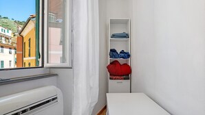 Une pièce avec une fenêtre, des murs blancs, un radiateur blanc, un banc blanc et une étagère blanche où sont pliés des vêtements, ainsi qu’un coussin rouge.