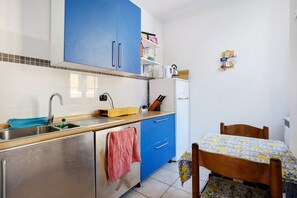 A compact kitchen with blue cabinets, a sink, a small refrigerator, and a dining table with a patterned tablecloth. - The 'Mira' Room with Shared Terrace, Wi-Fi and Air Conditioning (Vernazza)