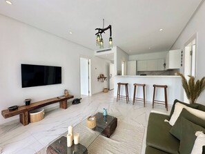 A modern living room with a flat-screen TV, a wooden bench, a glass coffee table with candles, a green sofa, and a kitchen area with white cabinets and a bar stool. - Appartement Serena Beachfront Hammamet (Hammamet)