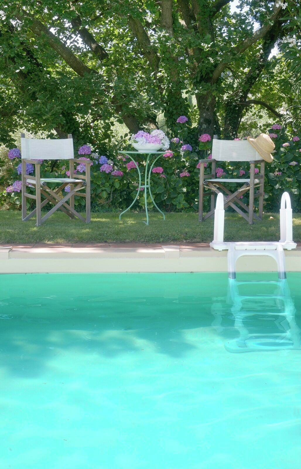 A pool area with two wooden chairs, a small table with a bowl of flowers, and a white pool ladder.