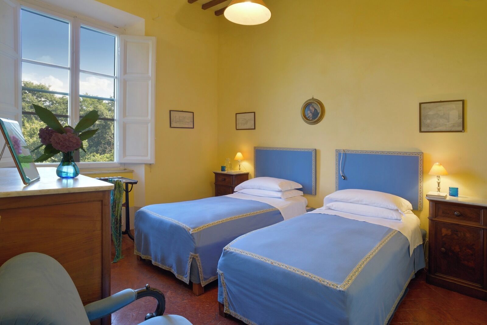 A room with two beds, a desk with a vase of flowers, and a window with a view of trees.