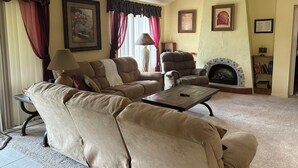 Een woonkamer met een bank, fauteuil, open haard en plafondventilator.