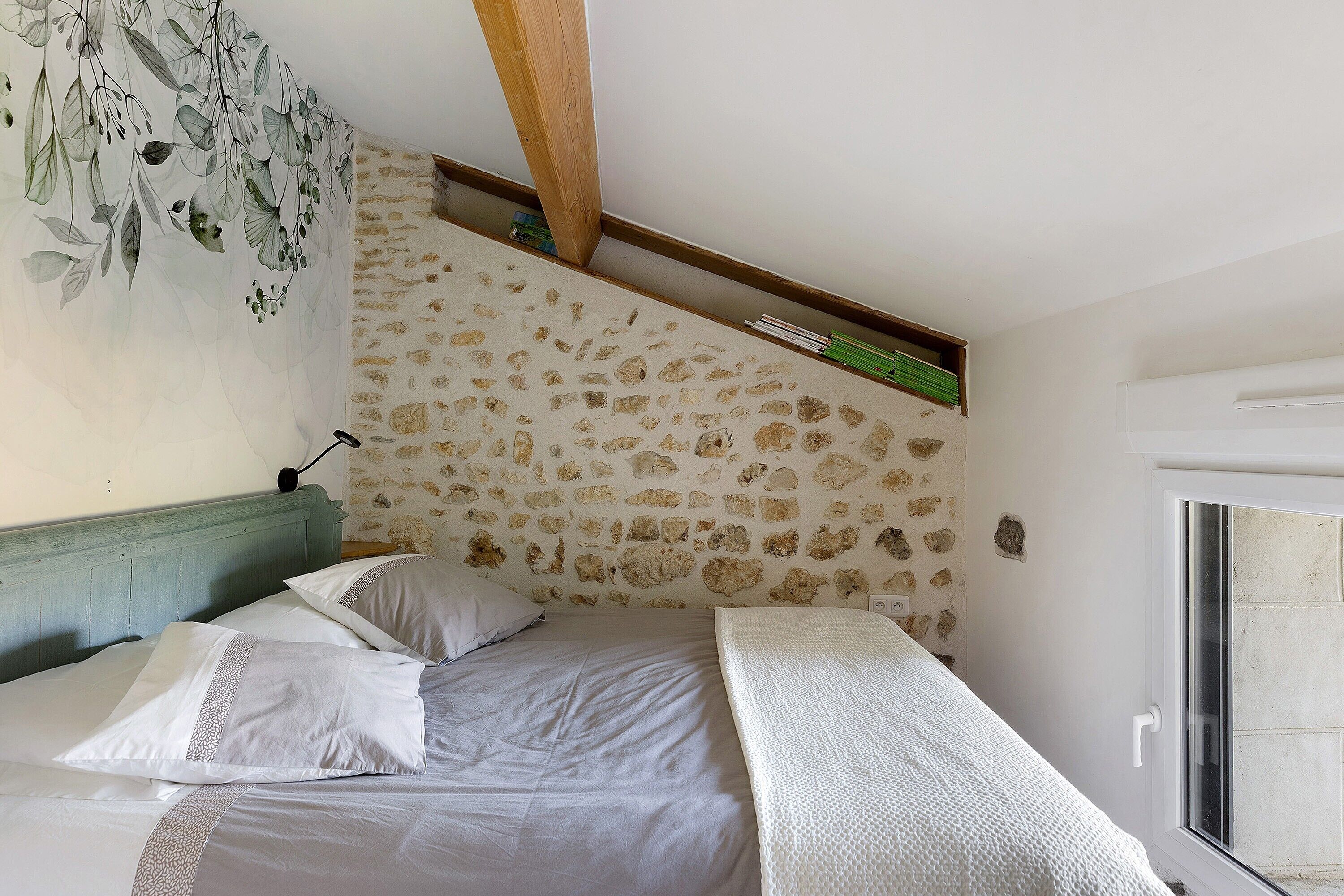 Camera da letto con parete in pietra, trave di legno al soffitto e un letto con biancheria bianca, oltre a una finestra.