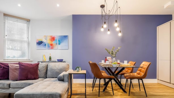 Una sala de estar moderna con un sofá gris, una mesa de comedor con cuatro sillas y un cuadro abstracto y colorido en la pared.