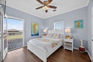 A bedroom with a bed, ceiling fan, artwork, bedside tables, and a view of a deck. - King Crab - Pet Friendly, Steps to the Beach! (Crystal Beach)