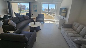 Ein modernes Wohnzimmer mit einer Couch, Sesseln, einem Couchtisch und einem Fernseher. Große Fenster bieten Blick auf Gebäude und einen wolkenlosen Himmel.