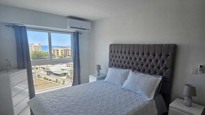 Ein Schlafzimmer mit einem großen Bett, einem Nachttisch, einer Kommode und einem Fenster mit Blick auf Gebäude und ein Gewässer.