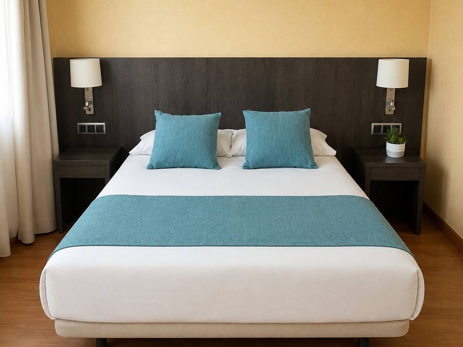 A hotel room with a bed, two nightstands, a lamp on each nightstand, a plant on one nightstand, and a window with curtains.