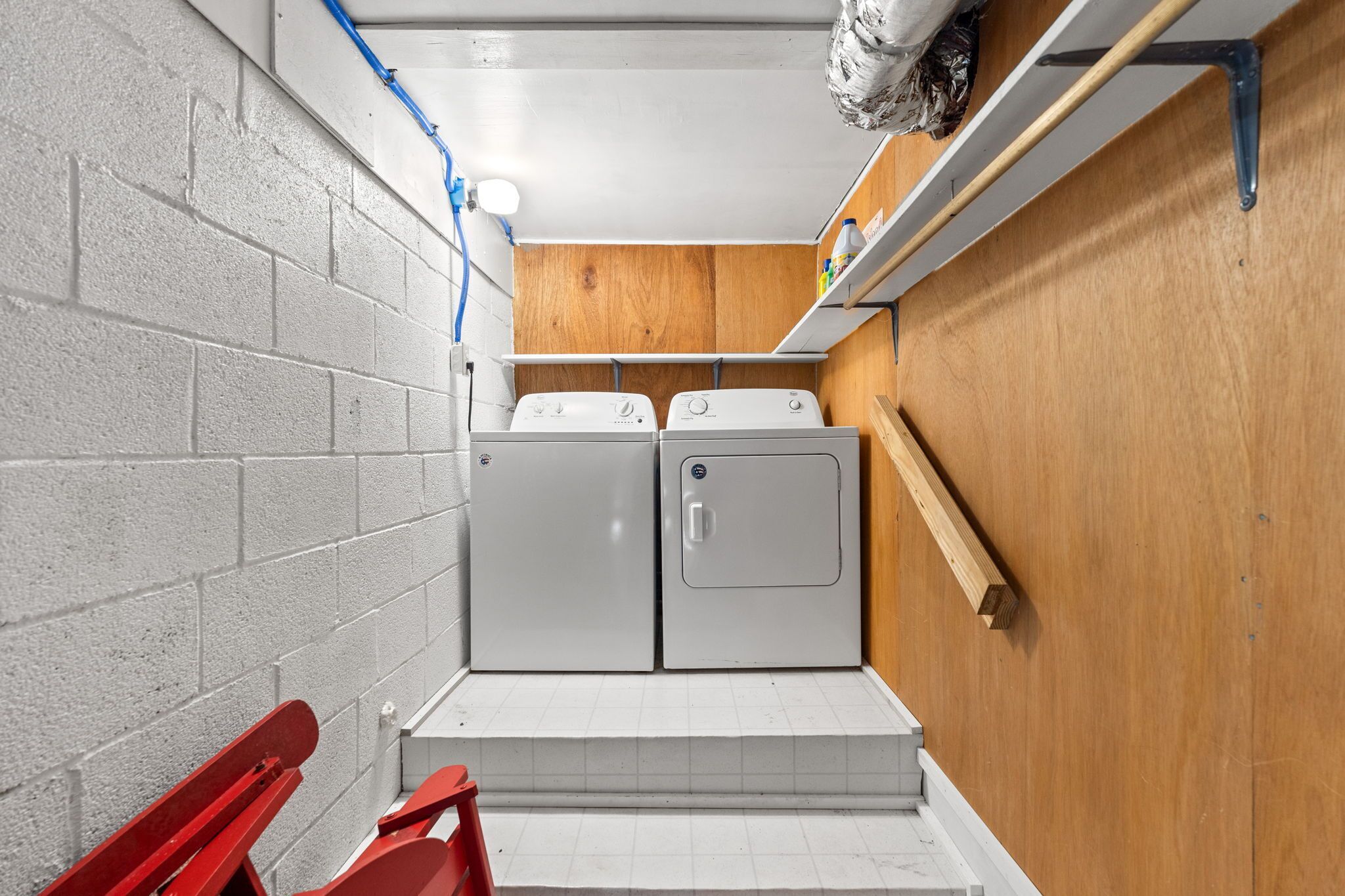 A laundry room with two white washers and dryers, a wooden shelf, and a red chair.