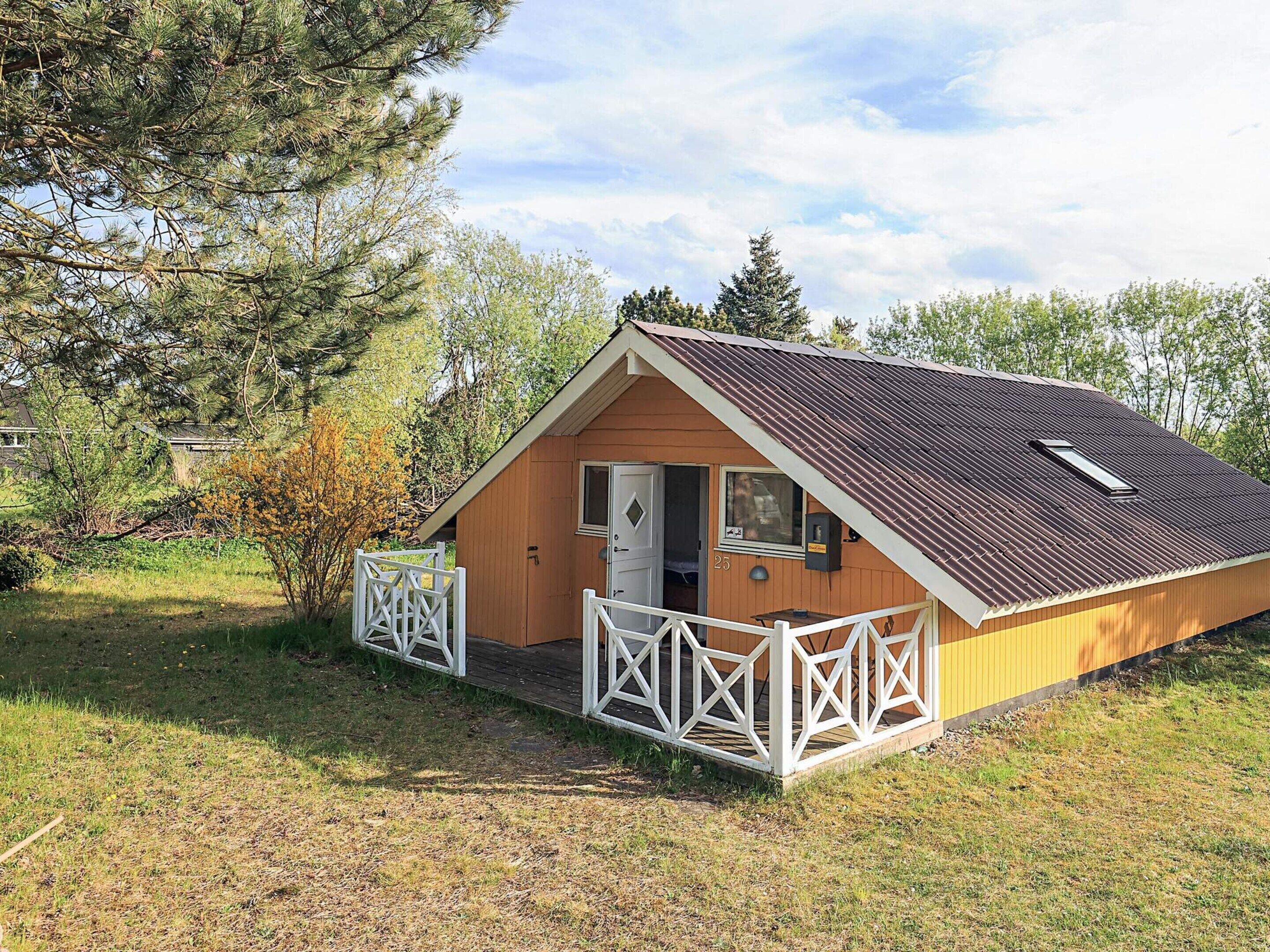 Une petite cabane en bois d’un seul étage, avec un toit en pente, une porte d’entrée et un petit porche doté d’une balustrade blanche.