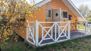 A wooden cabin with a white porch and a door, surrounded by trees with yellow flowers. - 5 Person Holiday Home in Nakskov (Nakskov)