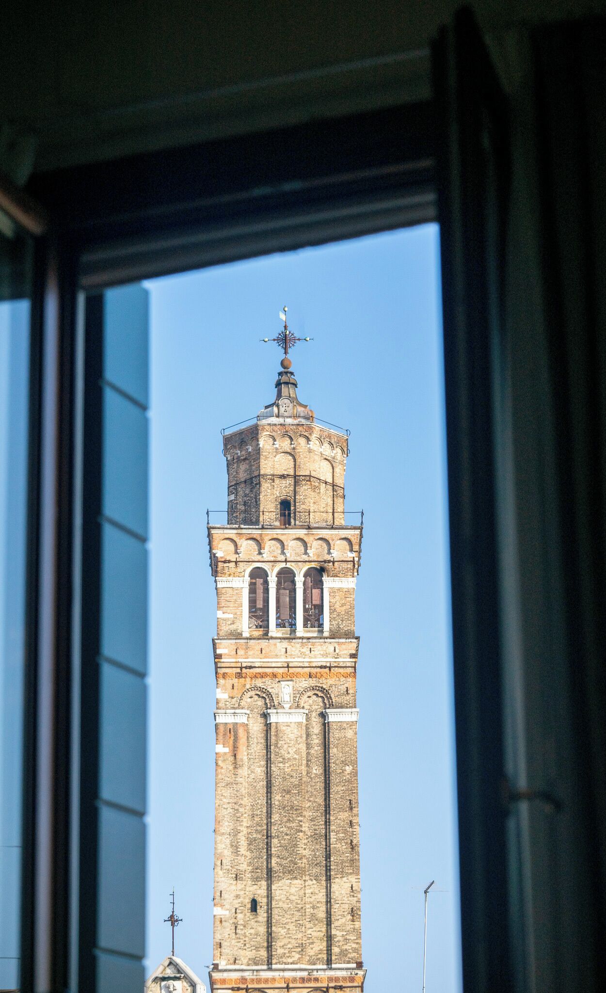 A tall brick tower with a weather vane, through a window with blinds.