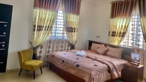 Un dormitorio con cama, una silla, una mesita de noche, un ventilador de techo y cortinas.
