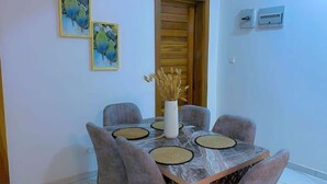 Un comedor con una mesa de mármol, cuatro sillas grises, un jarrón blanco con plantas secas y dos obras de arte enmarcadas en la pared.