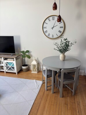 A room with a round table, a potted plant, a wall clock, a TV, and a small cabinet. - Comfortable Condo in Hoboken - 20 Minutes to MetLife Stadium for World Cup (Hoboken)