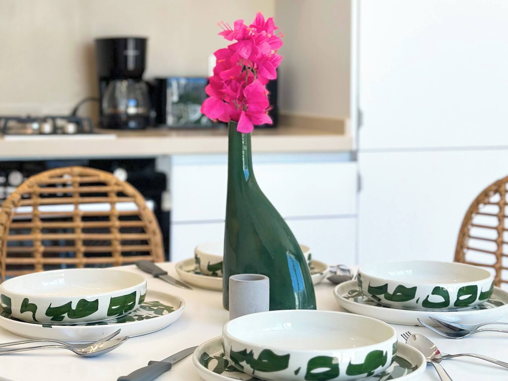 A dining table with green leaf-patterned bowls, a pink flower in a green vase, and a coffee machine in the background.