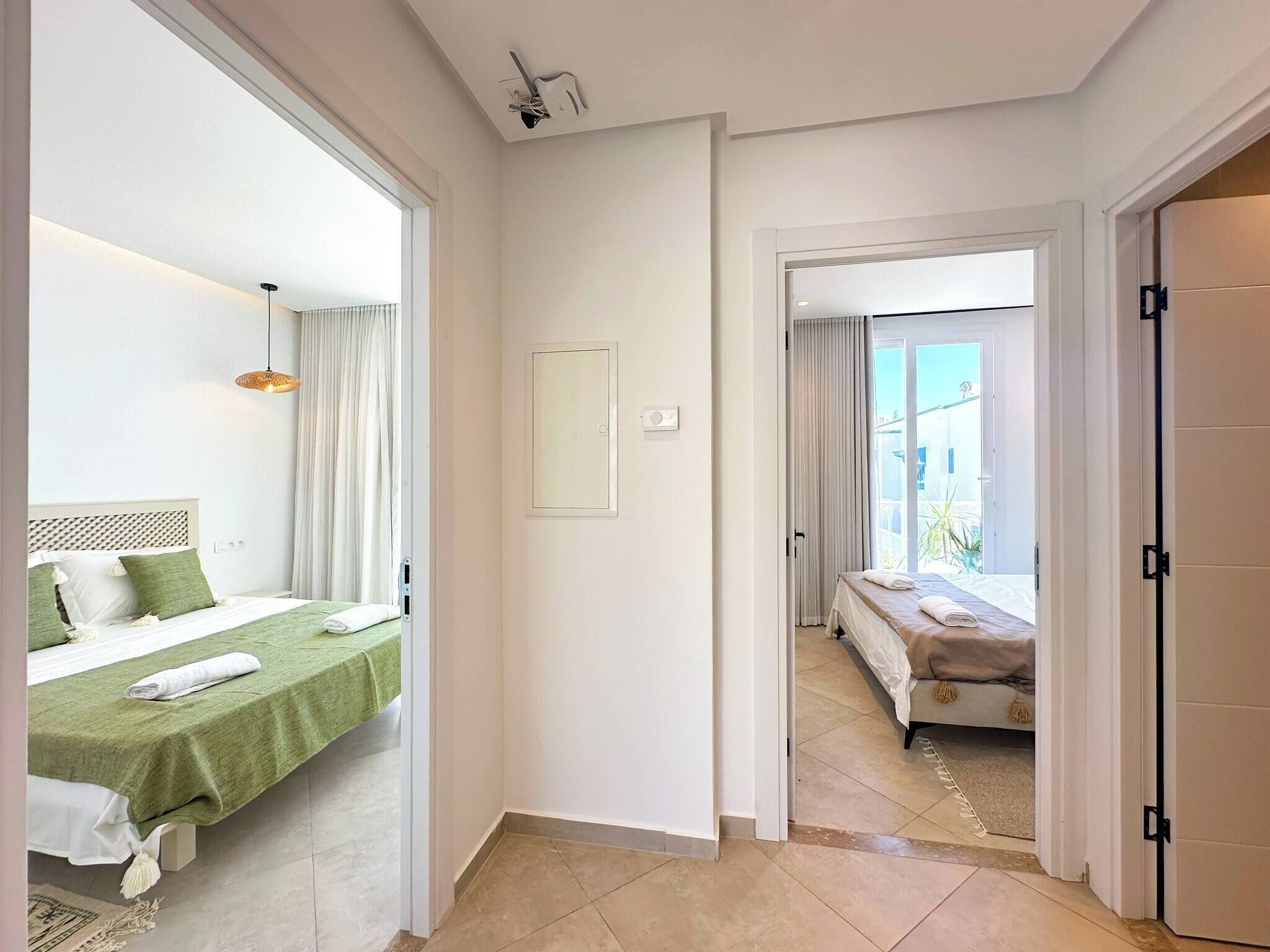 Hotellihuone, jossa on sänky, pieni pöytä, tuoli, ikkuna ja ovi, joka johtaa toiseen huoneeseen.