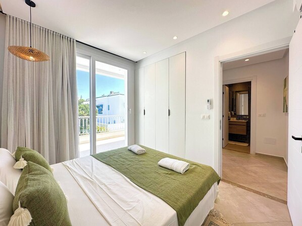 A hotel room with a bed, a green throw, a white towel, a balcony with curtains, and a view of the outside.