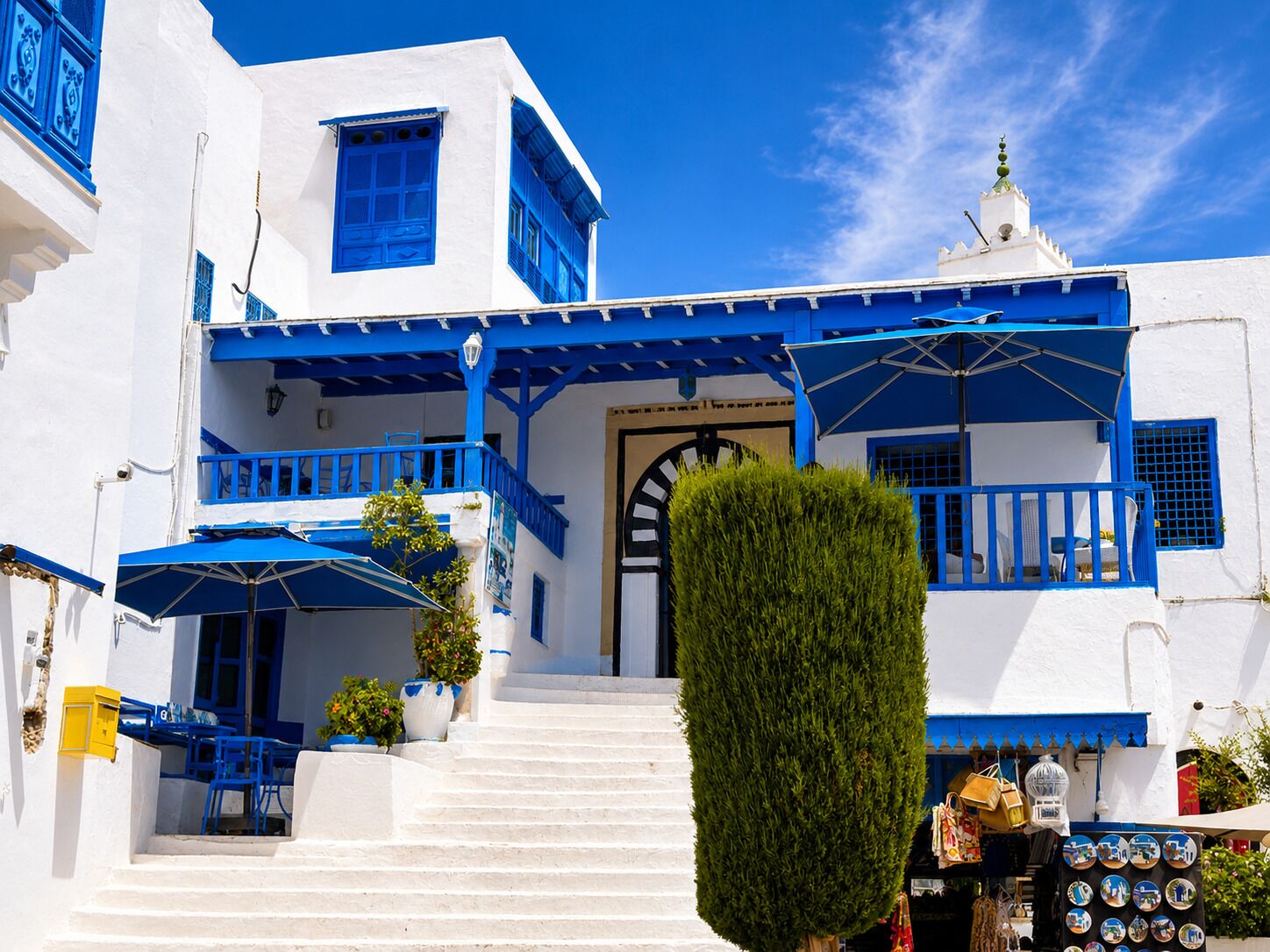 A white building with blue accents, a staircase leading to the entrance, and outdoor seating.