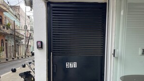 Une porte noire fermée, portant le numéro 3711, un mur blanc et une vue sur la rue, avec des immeubles et des scooters garés.