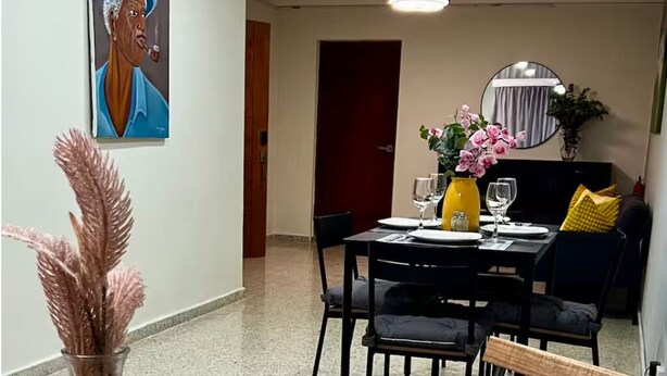 Un coin repas avec une table dressée pour quatre personnes, des chaises, un vase rempli de fleurs, un tableau accroché au mur et un ventilateur de plafond.