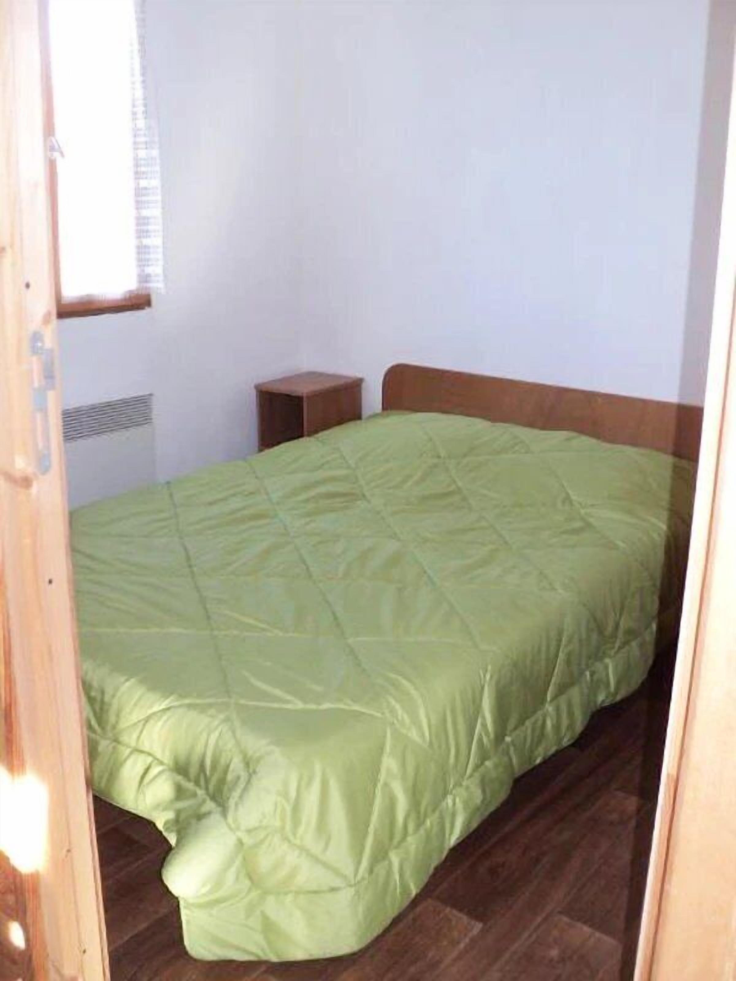 Una cama individual con cubierta verde en una habitación con paredes blancas y piso de madera.